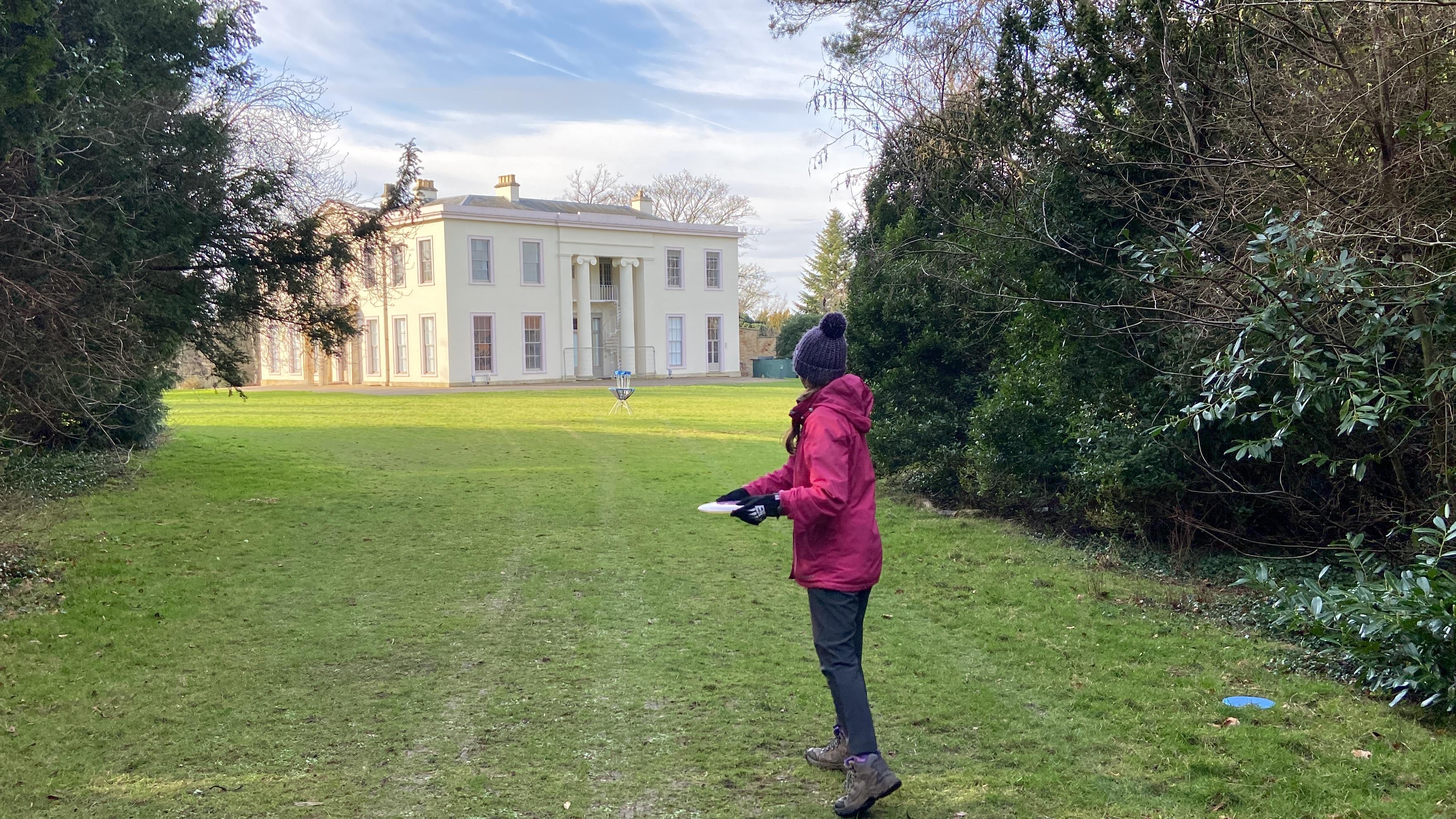 A disc golfer about to throw towards a basket with Papworth Hall in the background